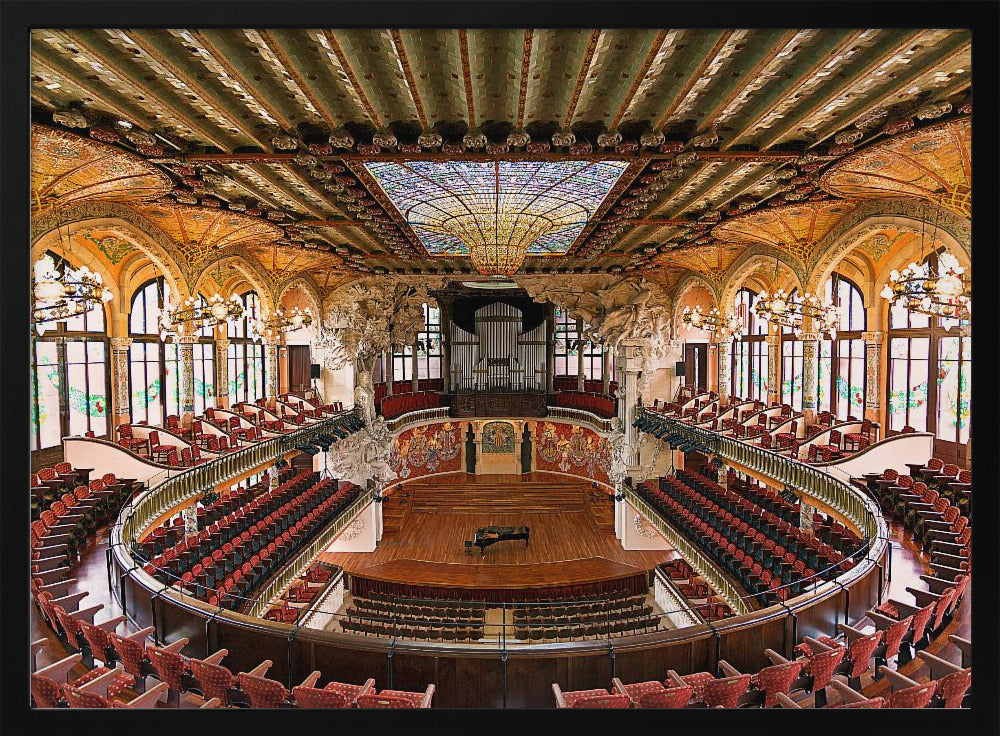 A high-angle, wide shot of the ornate and lavish interior of the Palau de la Música Catalana in Barcelona. The view captures the entire concert hall, with its curved balconies filled with red seats, a central wooden stage with a grand piano, and a spectacular, large stained-glass skylight in the ceiling. Poster