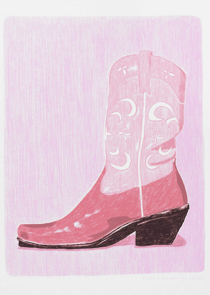 A framed art print of a pink cowboy boot against a light pink, textured background. The boot is illustrated in a block-print style with shades of pink, white decorative inlay patterns of moons and stars, and a dark brown heel and sole. Decor