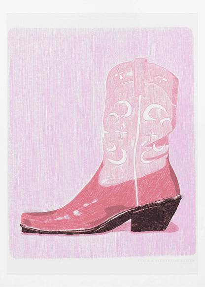 A framed art print of a pink cowboy boot against a light pink, textured background. The boot is illustrated in a block-print style with shades of pink, white decorative inlay patterns of moons and stars, and a dark brown heel and sole. Decor