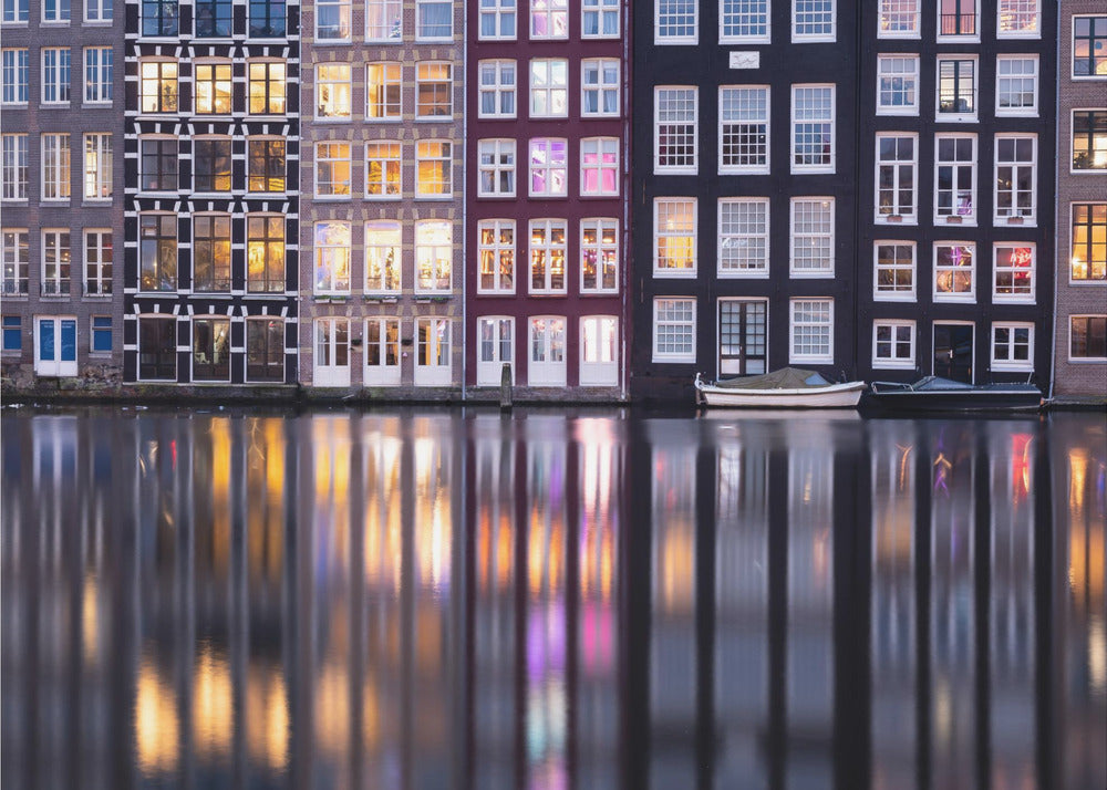A framed photograph of colorful Amsterdam canal houses at night, their illuminated windows casting vibrant, shimmering reflections on the water below. Artwork