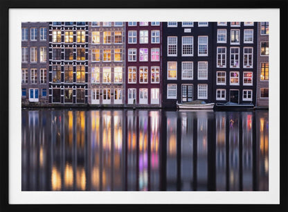 A framed photograph of colorful Amsterdam canal houses at night, their illuminated windows casting vibrant, shimmering reflections on the water below. Artwork