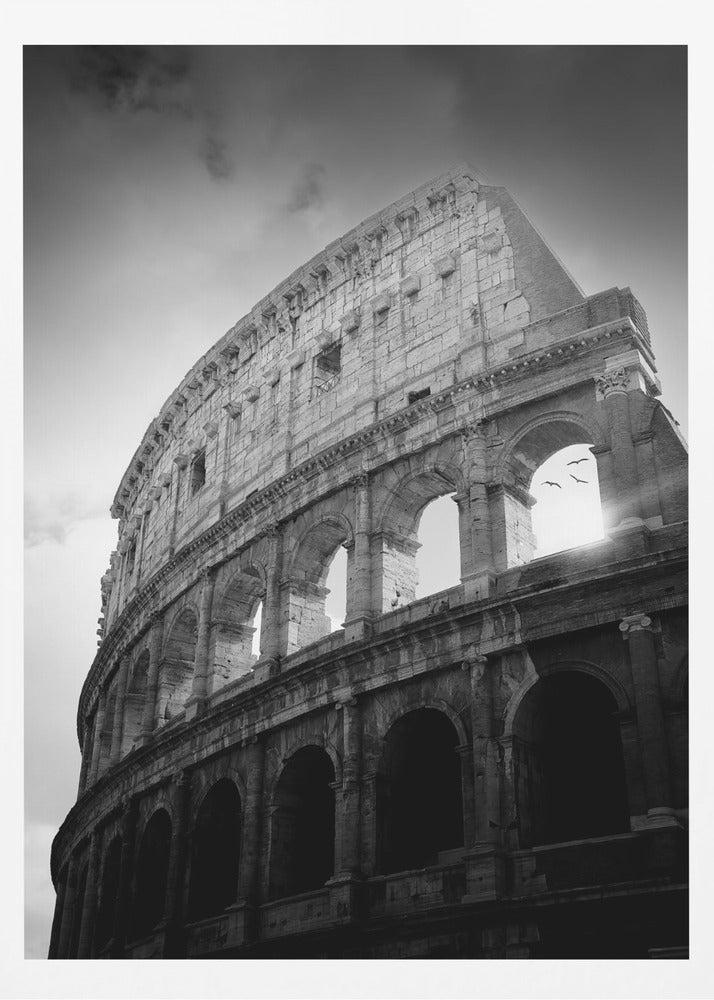 A dramatic, low-angle black and white photograph of the ancient Roman Colosseum. The sun shines brightly through one of the upper archways, creating a lens flare and silhouetting a few birds in flight against the cloudy sky. Poster