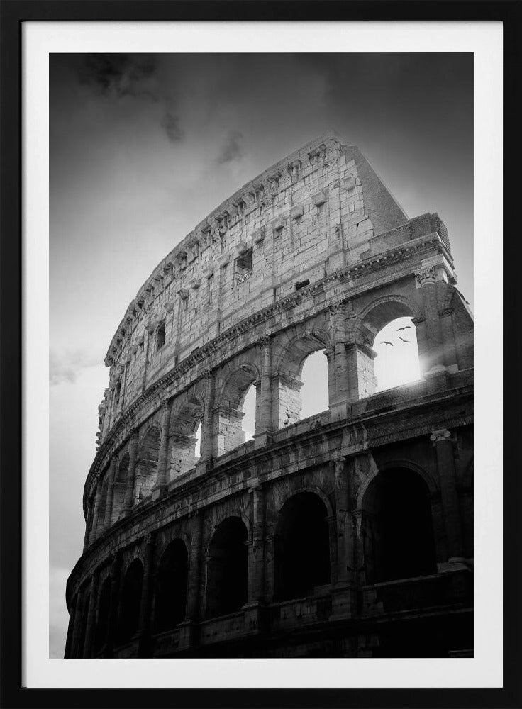 A dramatic, low-angle black and white photograph of the ancient Roman Colosseum. The sun shines brightly through one of the upper archways, creating a lens flare and silhouetting a few birds in flight against the cloudy sky. Poster