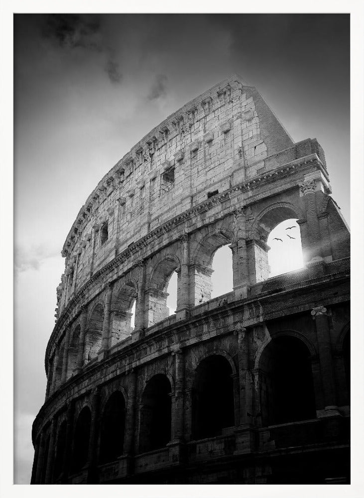 A dramatic, low-angle black and white photograph of the ancient Roman Colosseum. The sun shines brightly through one of the upper archways, creating a lens flare and silhouetting a few birds in flight against the cloudy sky. Poster