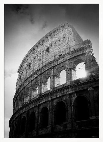 A dramatic, low-angle black and white photograph of the ancient Roman Colosseum. The sun shines brightly through one of the upper archways, creating a lens flare and silhouetting a few birds in flight against the cloudy sky. Poster