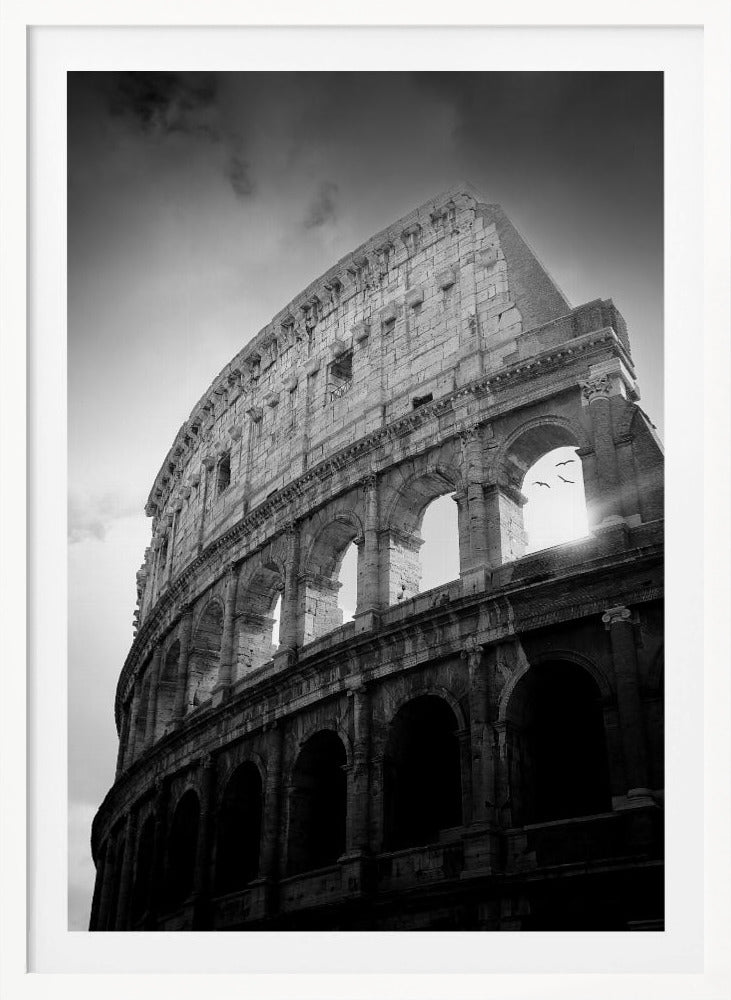 A dramatic, low-angle black and white photograph of the ancient Roman Colosseum. The sun shines brightly through one of the upper archways, creating a lens flare and silhouetting a few birds in flight against the cloudy sky. Poster