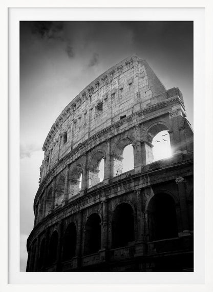 A dramatic, low-angle black and white photograph of the ancient Roman Colosseum. The sun shines brightly through one of the upper archways, creating a lens flare and silhouetting a few birds in flight against the cloudy sky. Poster