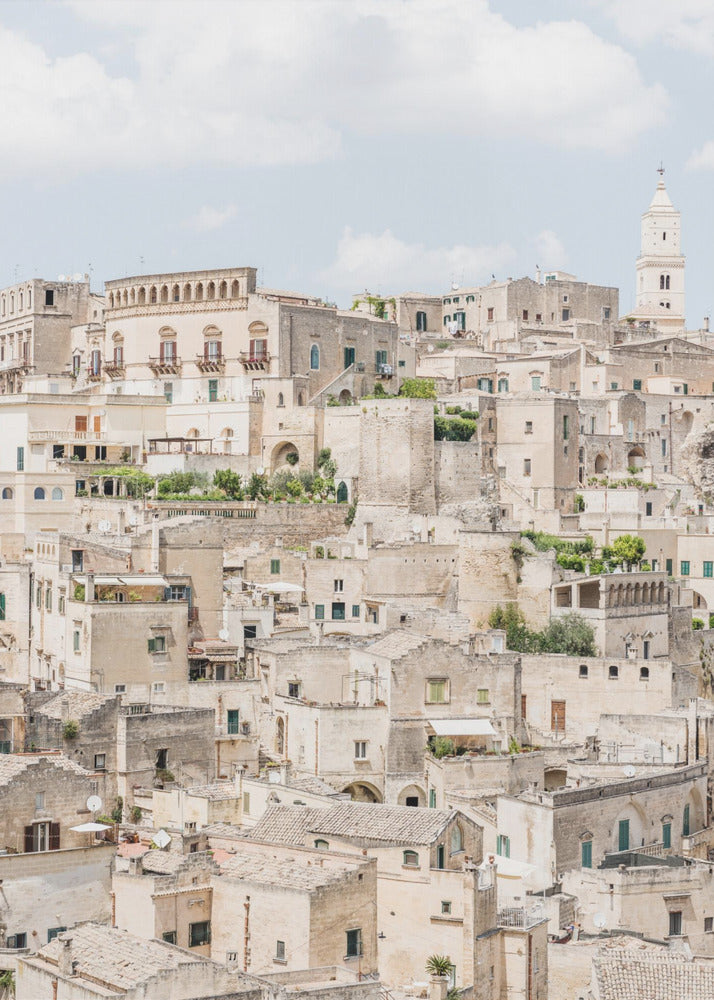 A vertical photograph of the ancient, stone city of Matera, Italy, with its dense collection of beige buildings and tiled roofs built up a hillside under a pale blue sky. A white bell tower stands out against the skyline. Poster