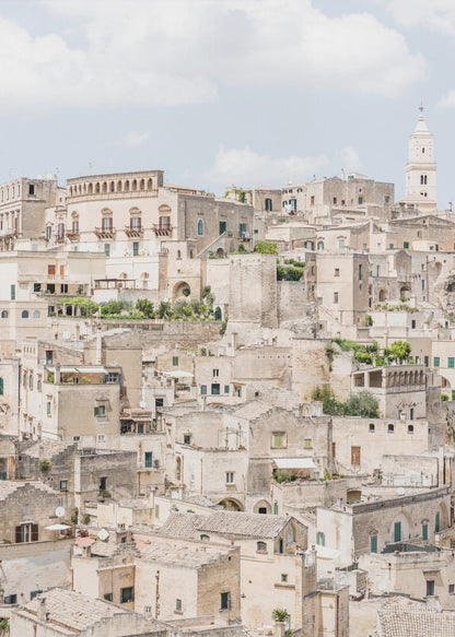 A vertical photograph of the ancient, stone city of Matera, Italy, with its dense collection of beige buildings and tiled roofs built up a hillside under a pale blue sky. A white bell tower stands out against the skyline. Poster