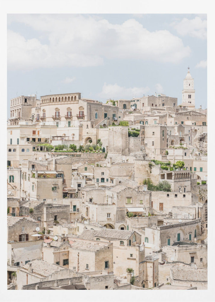 A vertical photograph of the ancient, stone city of Matera, Italy, with its dense collection of beige buildings and tiled roofs built up a hillside under a pale blue sky. A white bell tower stands out against the skyline. Poster