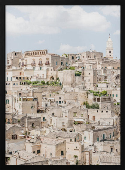 A vertical photograph of the ancient, stone city of Matera, Italy, with its dense collection of beige buildings and tiled roofs built up a hillside under a pale blue sky. A white bell tower stands out against the skyline. Poster
