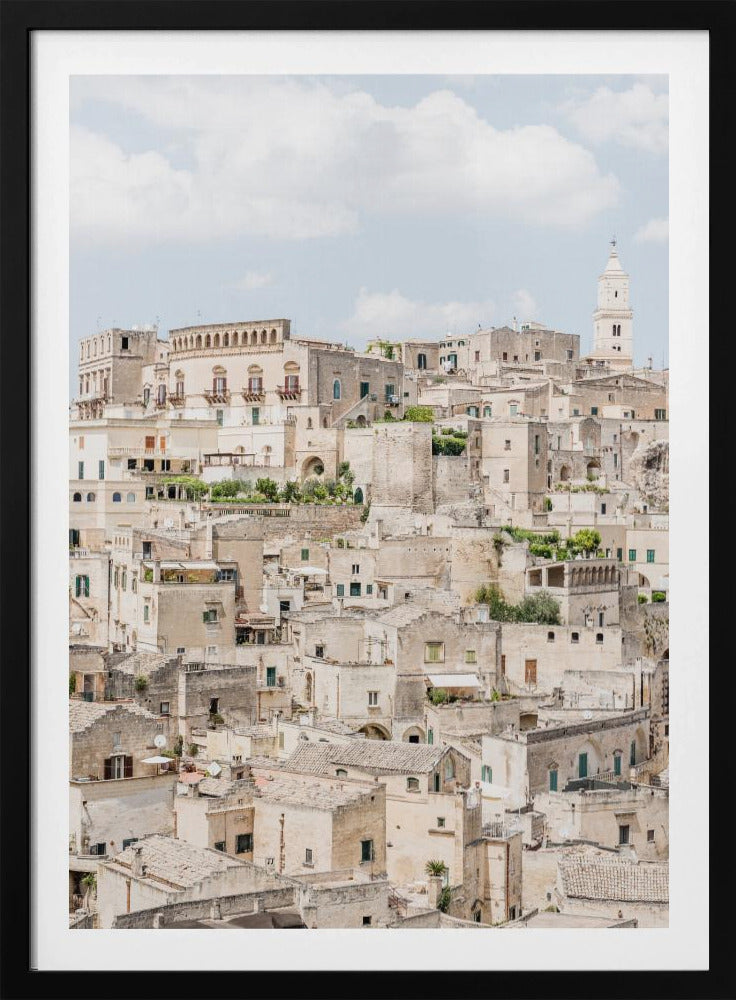 A vertical photograph of the ancient, stone city of Matera, Italy, with its dense collection of beige buildings and tiled roofs built up a hillside under a pale blue sky. A white bell tower stands out against the skyline. Poster