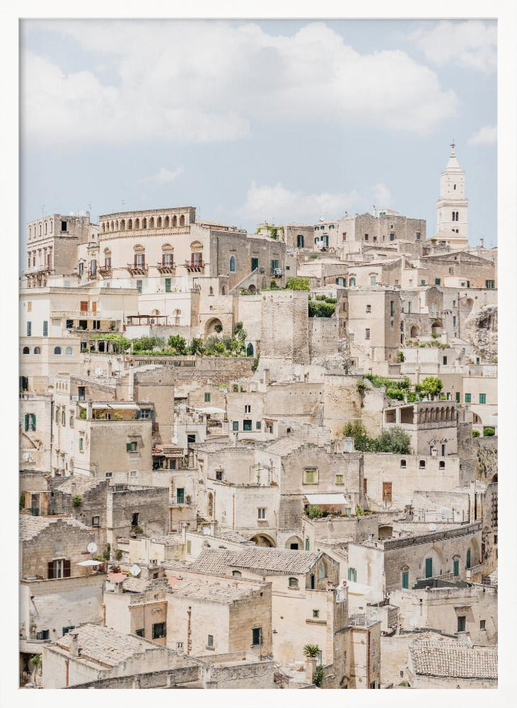 A vertical photograph of the ancient, stone city of Matera, Italy, with its dense collection of beige buildings and tiled roofs built up a hillside under a pale blue sky. A white bell tower stands out against the skyline. Poster