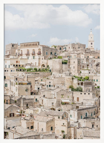 A vertical photograph of the ancient, stone city of Matera, Italy, with its dense collection of beige buildings and tiled roofs built up a hillside under a pale blue sky. A white bell tower stands out against the skyline. Poster