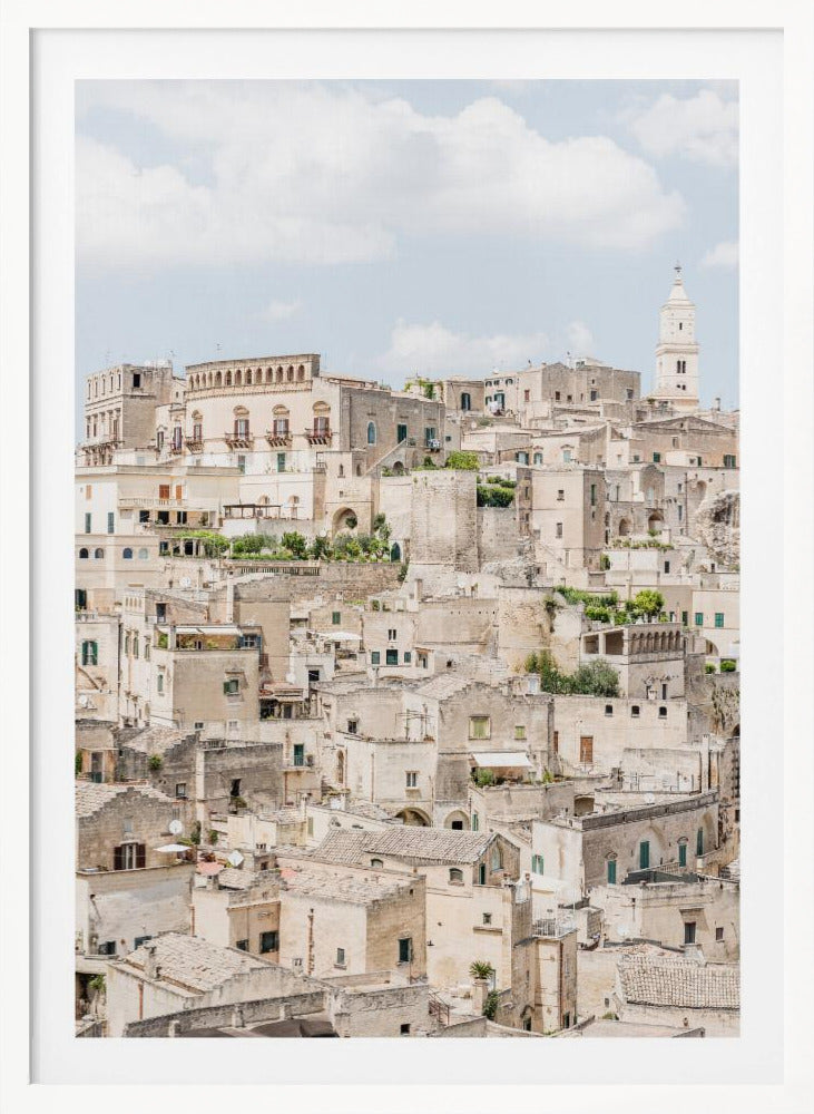 A vertical photograph of the ancient, stone city of Matera, Italy, with its dense collection of beige buildings and tiled roofs built up a hillside under a pale blue sky. A white bell tower stands out against the skyline. Poster
