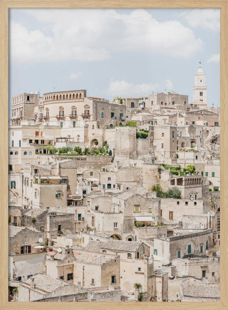 A vertical photograph of the ancient, stone city of Matera, Italy, with its dense collection of beige buildings and tiled roofs built up a hillside under a pale blue sky. A white bell tower stands out against the skyline. Poster
