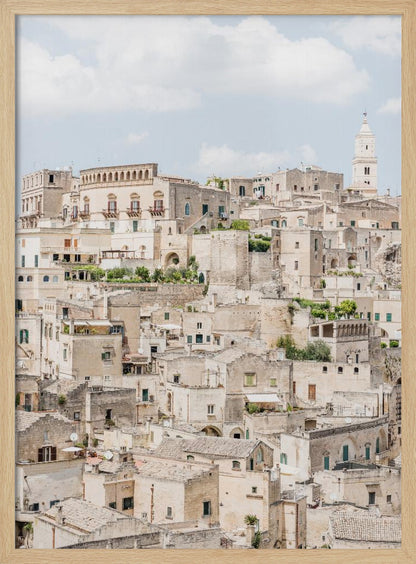 A vertical photograph of the ancient, stone city of Matera, Italy, with its dense collection of beige buildings and tiled roofs built up a hillside under a pale blue sky. A white bell tower stands out against the skyline. Poster