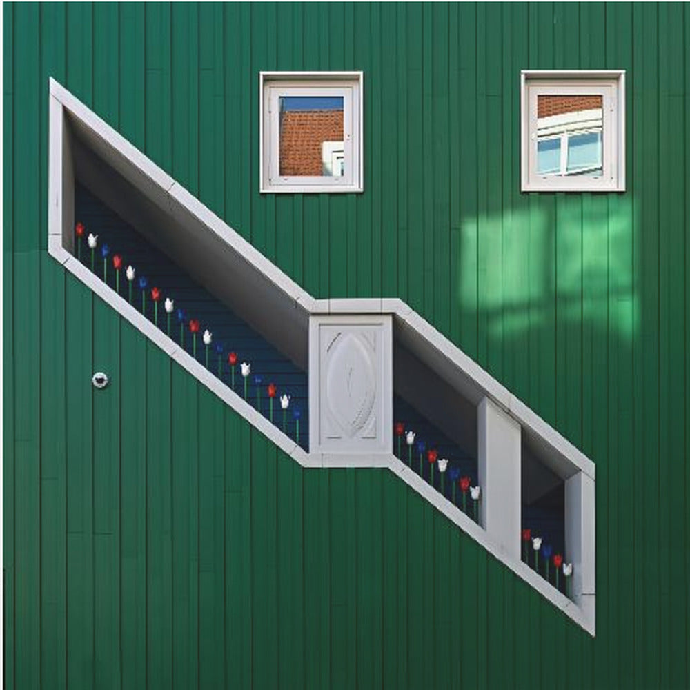 A framed photograph of a modern building with a dark green vertical-paneled facade, featuring a large, white-framed diagonal window that resembles a staircase, decorated with small red, white, and blue tulips. Two small square windows are positioned above. Decor