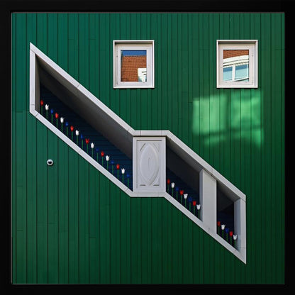 A framed photograph of a modern building with a dark green vertical-paneled facade, featuring a large, white-framed diagonal window that resembles a staircase, decorated with small red, white, and blue tulips. Two small square windows are positioned above. Decor