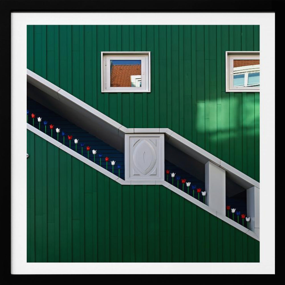 A framed photograph of a modern building with a dark green vertical-paneled facade, featuring a large, white-framed diagonal window that resembles a staircase, decorated with small red, white, and blue tulips. Two small square windows are positioned above. Decor