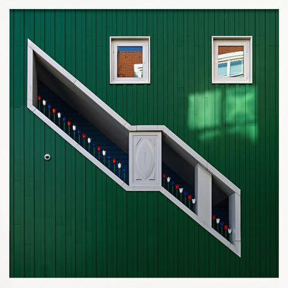 A framed photograph of a modern building with a dark green vertical-paneled facade, featuring a large, white-framed diagonal window that resembles a staircase, decorated with small red, white, and blue tulips. Two small square windows are positioned above. Decor