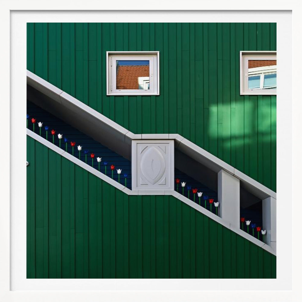A framed photograph of a modern building with a dark green vertical-paneled facade, featuring a large, white-framed diagonal window that resembles a staircase, decorated with small red, white, and blue tulips. Two small square windows are positioned above. Decor