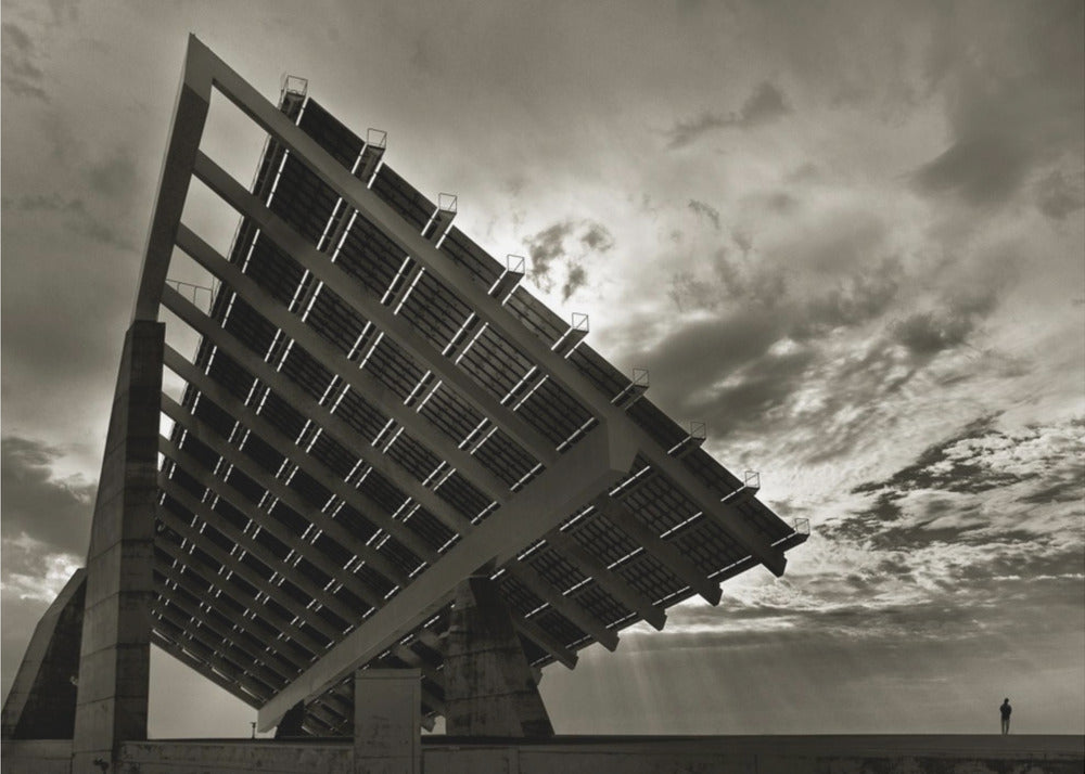 A framed, dramatic black and white photograph showing a massive, tilted solar panel structure from a low angle. The sky is filled with dark, moody clouds with some sun rays breaking through. A tiny silhouette of a person in the lower right corner emphasizes the grand scale of the architecture. Wall Art