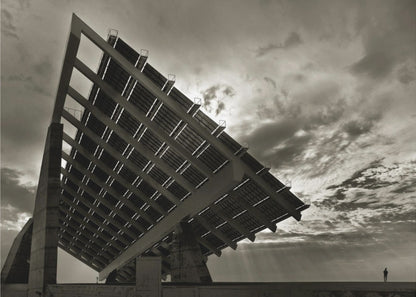 A framed, dramatic black and white photograph showing a massive, tilted solar panel structure from a low angle. The sky is filled with dark, moody clouds with some sun rays breaking through. A tiny silhouette of a person in the lower right corner emphasizes the grand scale of the architecture. Wall Art