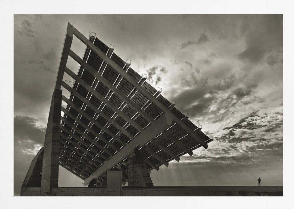A framed, dramatic black and white photograph showing a massive, tilted solar panel structure from a low angle. The sky is filled with dark, moody clouds with some sun rays breaking through. A tiny silhouette of a person in the lower right corner emphasizes the grand scale of the architecture. Wall Art