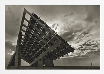 A framed, dramatic black and white photograph showing a massive, tilted solar panel structure from a low angle. The sky is filled with dark, moody clouds with some sun rays breaking through. A tiny silhouette of a person in the lower right corner emphasizes the grand scale of the architecture. Wall Art