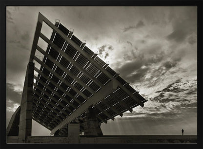 A framed, dramatic black and white photograph showing a massive, tilted solar panel structure from a low angle. The sky is filled with dark, moody clouds with some sun rays breaking through. A tiny silhouette of a person in the lower right corner emphasizes the grand scale of the architecture. Wall Art