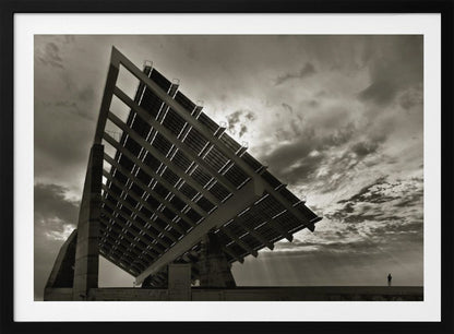 A framed, dramatic black and white photograph showing a massive, tilted solar panel structure from a low angle. The sky is filled with dark, moody clouds with some sun rays breaking through. A tiny silhouette of a person in the lower right corner emphasizes the grand scale of the architecture. Wall Art