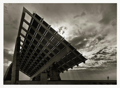 A framed, dramatic black and white photograph showing a massive, tilted solar panel structure from a low angle. The sky is filled with dark, moody clouds with some sun rays breaking through. A tiny silhouette of a person in the lower right corner emphasizes the grand scale of the architecture. Wall Art