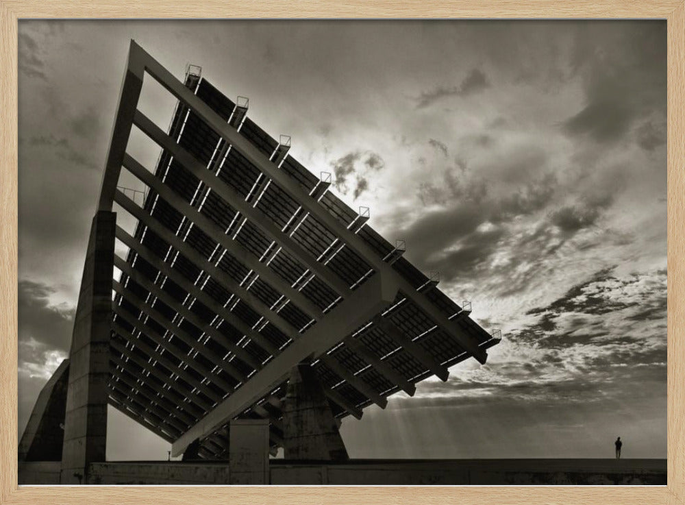 A framed, dramatic black and white photograph showing a massive, tilted solar panel structure from a low angle. The sky is filled with dark, moody clouds with some sun rays breaking through. A tiny silhouette of a person in the lower right corner emphasizes the grand scale of the architecture. Wall Art