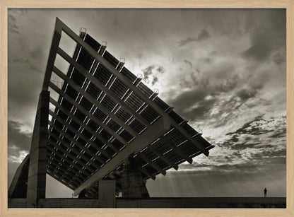 A framed, dramatic black and white photograph showing a massive, tilted solar panel structure from a low angle. The sky is filled with dark, moody clouds with some sun rays breaking through. A tiny silhouette of a person in the lower right corner emphasizes the grand scale of the architecture. Wall Art