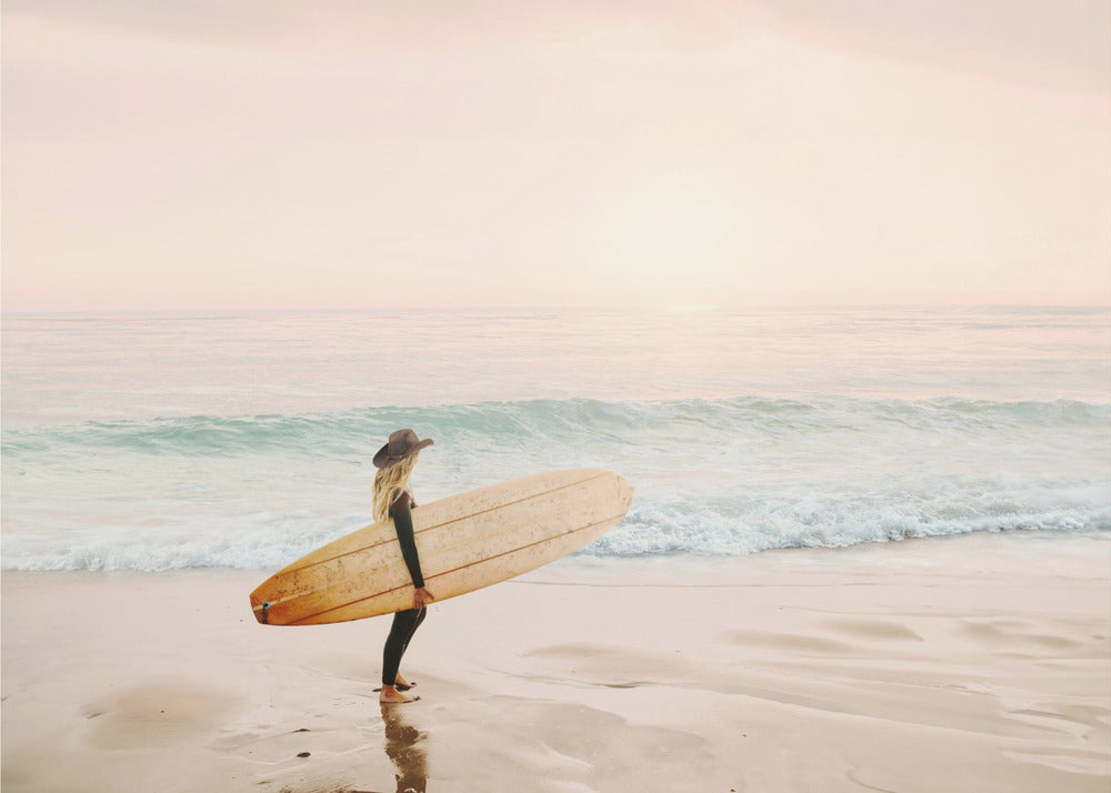 A female surfer with long blonde hair, wearing a black wetsuit and a hat, holds a surfboard while standing on a wet sandy beach looking out at the ocean during a hazy pink sunset. Decor