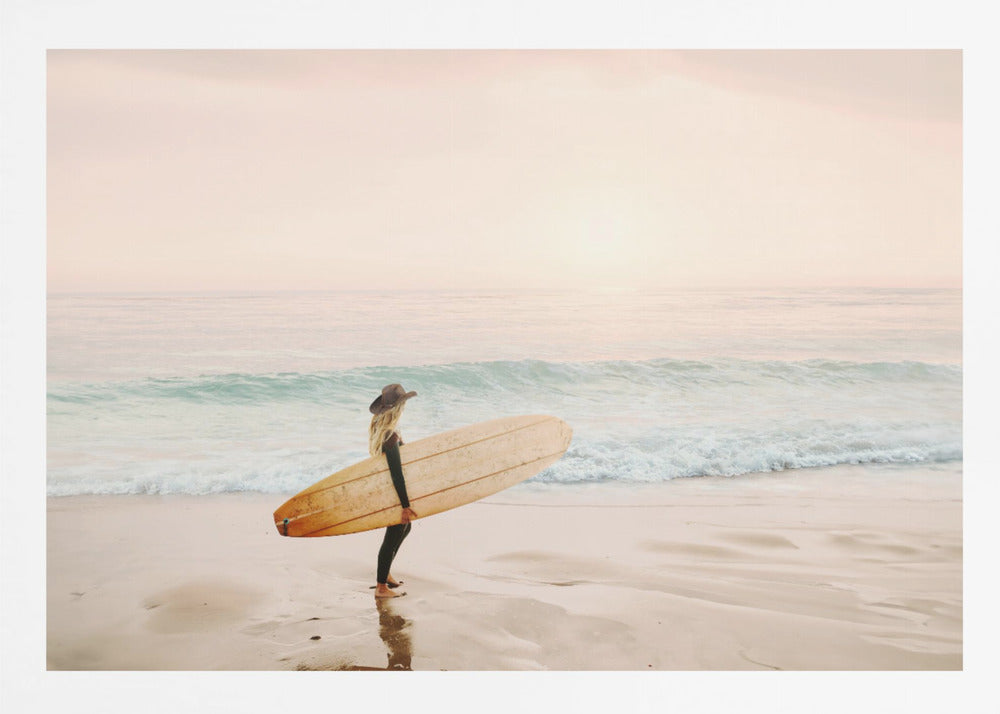 A female surfer with long blonde hair, wearing a black wetsuit and a hat, holds a surfboard while standing on a wet sandy beach looking out at the ocean during a hazy pink sunset. Decor