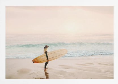 A female surfer with long blonde hair, wearing a black wetsuit and a hat, holds a surfboard while standing on a wet sandy beach looking out at the ocean during a hazy pink sunset. Decor