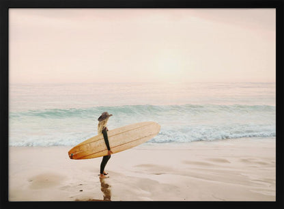 A female surfer with long blonde hair, wearing a black wetsuit and a hat, holds a surfboard while standing on a wet sandy beach looking out at the ocean during a hazy pink sunset. Decor