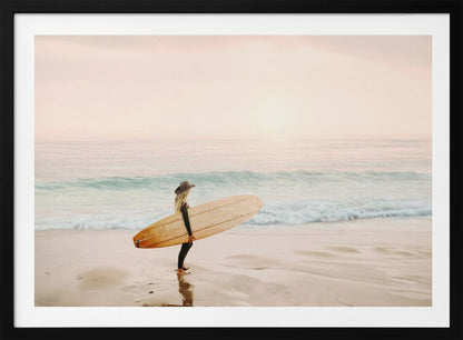 A female surfer with long blonde hair, wearing a black wetsuit and a hat, holds a surfboard while standing on a wet sandy beach looking out at the ocean during a hazy pink sunset. Decor