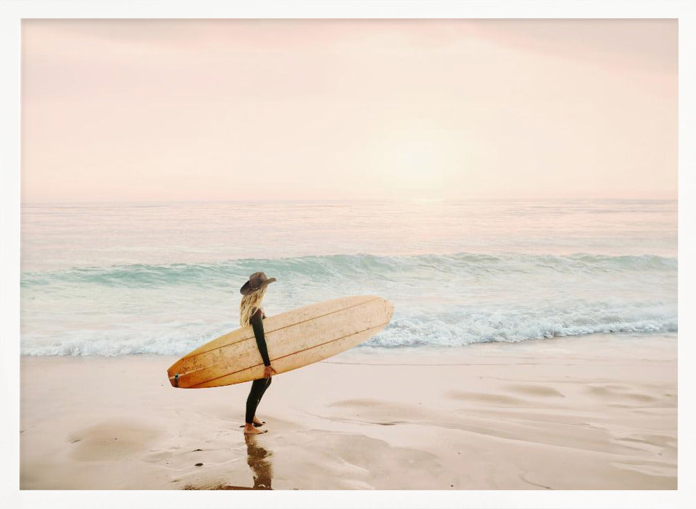 A female surfer with long blonde hair, wearing a black wetsuit and a hat, holds a surfboard while standing on a wet sandy beach looking out at the ocean during a hazy pink sunset. Decor