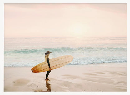 A female surfer with long blonde hair, wearing a black wetsuit and a hat, holds a surfboard while standing on a wet sandy beach looking out at the ocean during a hazy pink sunset. Decor