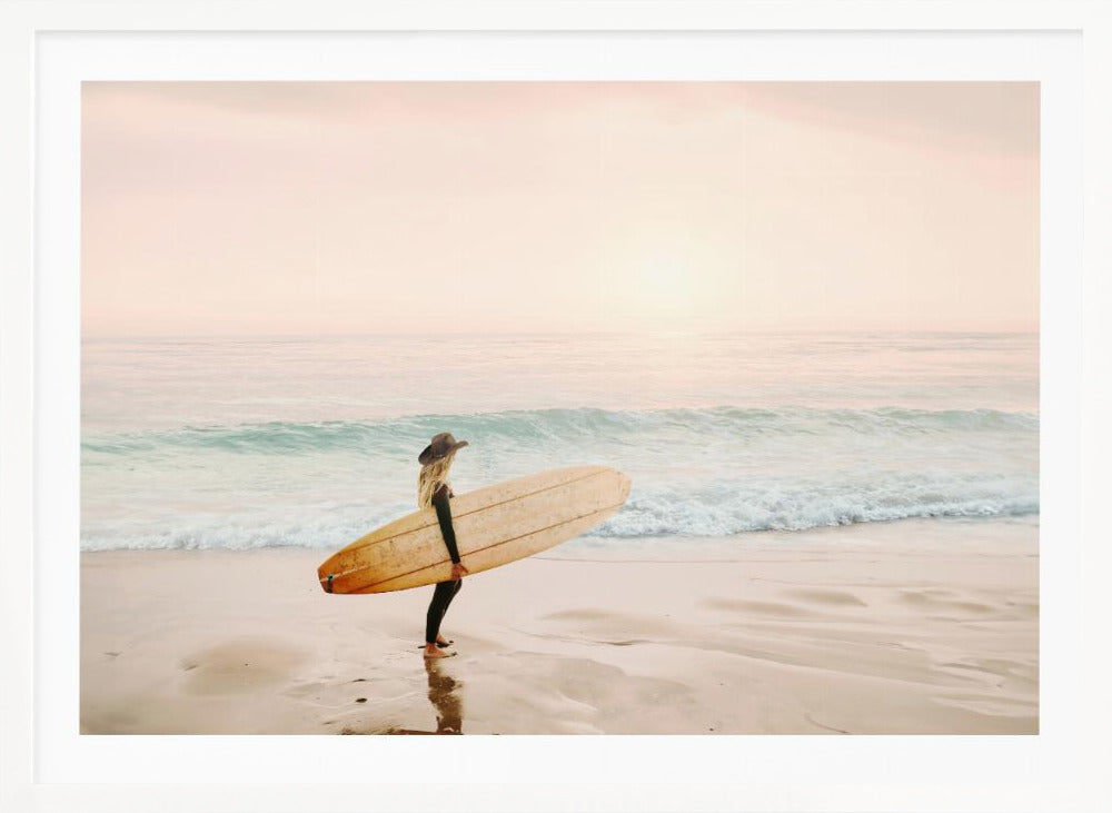 A female surfer with long blonde hair, wearing a black wetsuit and a hat, holds a surfboard while standing on a wet sandy beach looking out at the ocean during a hazy pink sunset. Decor