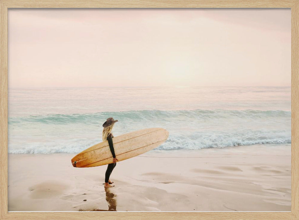 A female surfer with long blonde hair, wearing a black wetsuit and a hat, holds a surfboard while standing on a wet sandy beach looking out at the ocean during a hazy pink sunset. Decor
