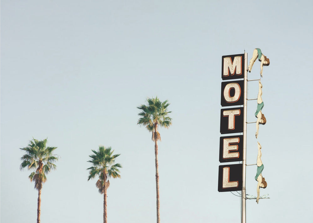 A minimalist photograph featuring three tall palm trees and a vertical black and white 'MOTEL' sign against a pale blue sky. Affixed to the sign's pole are several figures of women in green swimsuits, posed as if diving in a synchronized formation. Poster