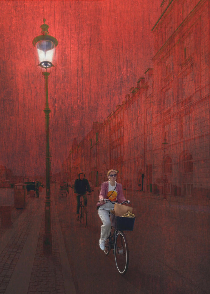 A surreal artistic image of a city street bathed in a textured red light. A woman rides a bicycle with a basket towards the viewer, while another cyclist is in the background. A tall, ornate lamppost is lit on the left side of the cobblestone street. Wall Art