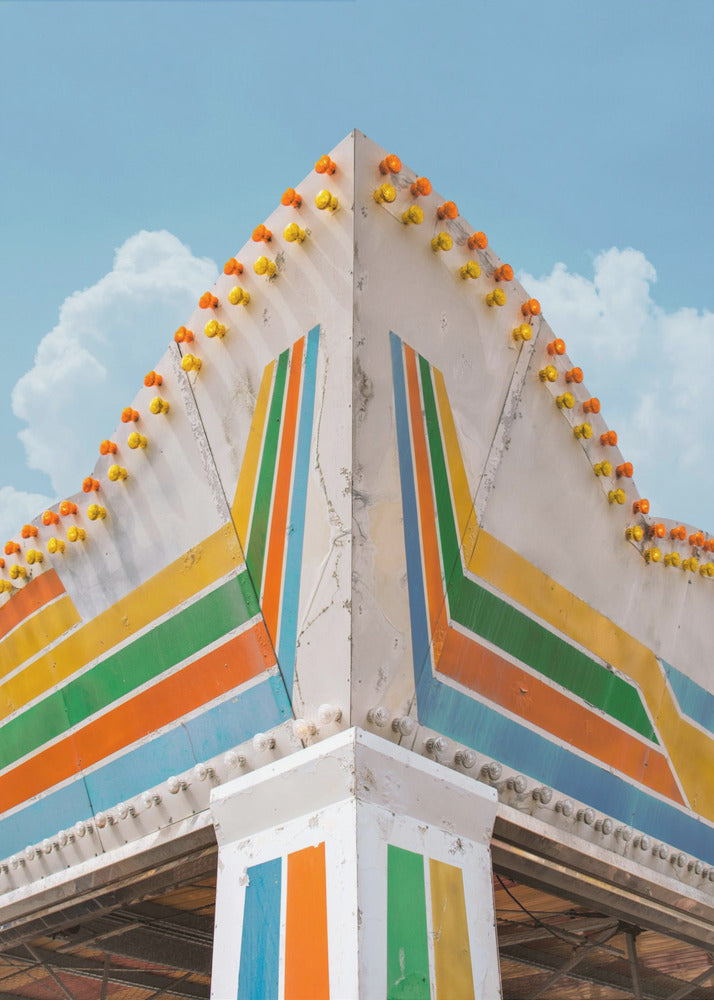 A low-angle photograph of the corner of a retro-style building against a bright blue sky with white clouds. The white facade is painted with bold, colorful stripes in orange, yellow, green, and blue, and the angled roofline is decorated with rows of unlit yellow and orange carnival lights. Print