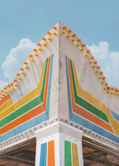 A low-angle photograph of the corner of a retro-style building against a bright blue sky with white clouds. The white facade is painted with bold, colorful stripes in orange, yellow, green, and blue, and the angled roofline is decorated with rows of unlit yellow and orange carnival lights. Print