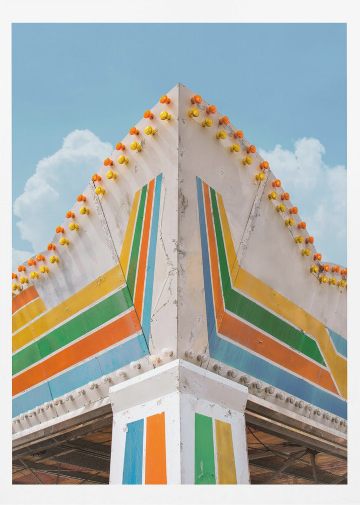 A low-angle photograph of the corner of a retro-style building against a bright blue sky with white clouds. The white facade is painted with bold, colorful stripes in orange, yellow, green, and blue, and the angled roofline is decorated with rows of unlit yellow and orange carnival lights. Print