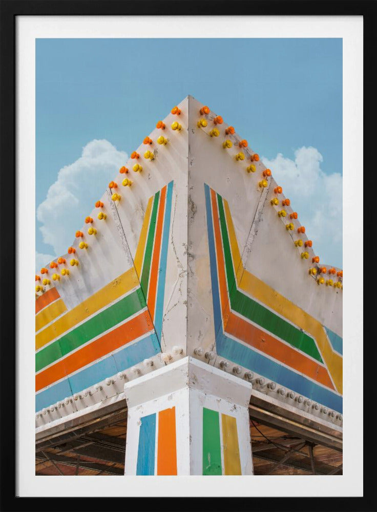 A low-angle photograph of the corner of a retro-style building against a bright blue sky with white clouds. The white facade is painted with bold, colorful stripes in orange, yellow, green, and blue, and the angled roofline is decorated with rows of unlit yellow and orange carnival lights. Print